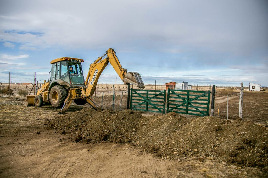 A yellow backhoe loader actively digs soil at a farm entrance with a green gate, under a cloudy sky.